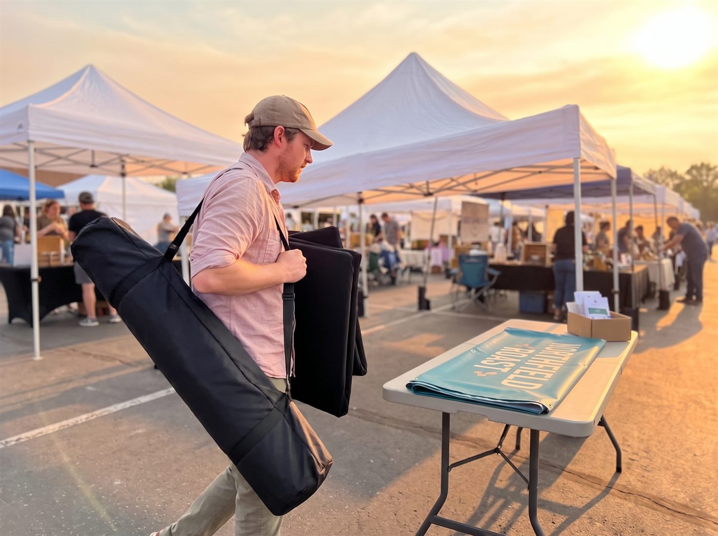 The vendor packing down at the end of the day, the retractable banner in its padded carry bag over one shoulder and both folded table covers under the other arm