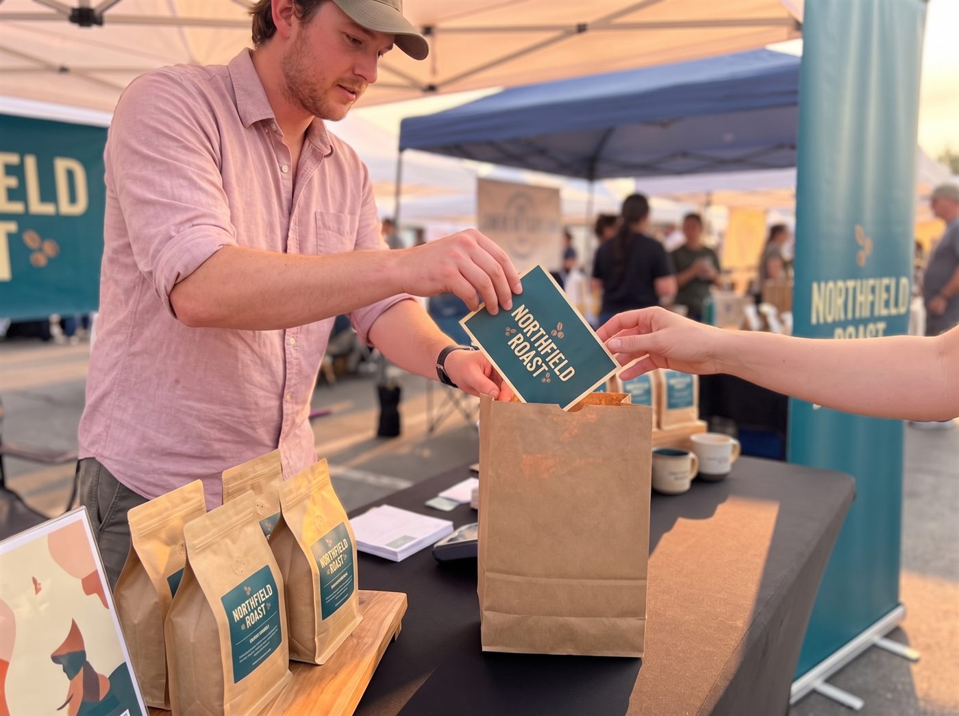 The vendor slipping a printed postcard into a kraft paper bag as a customer takes it
