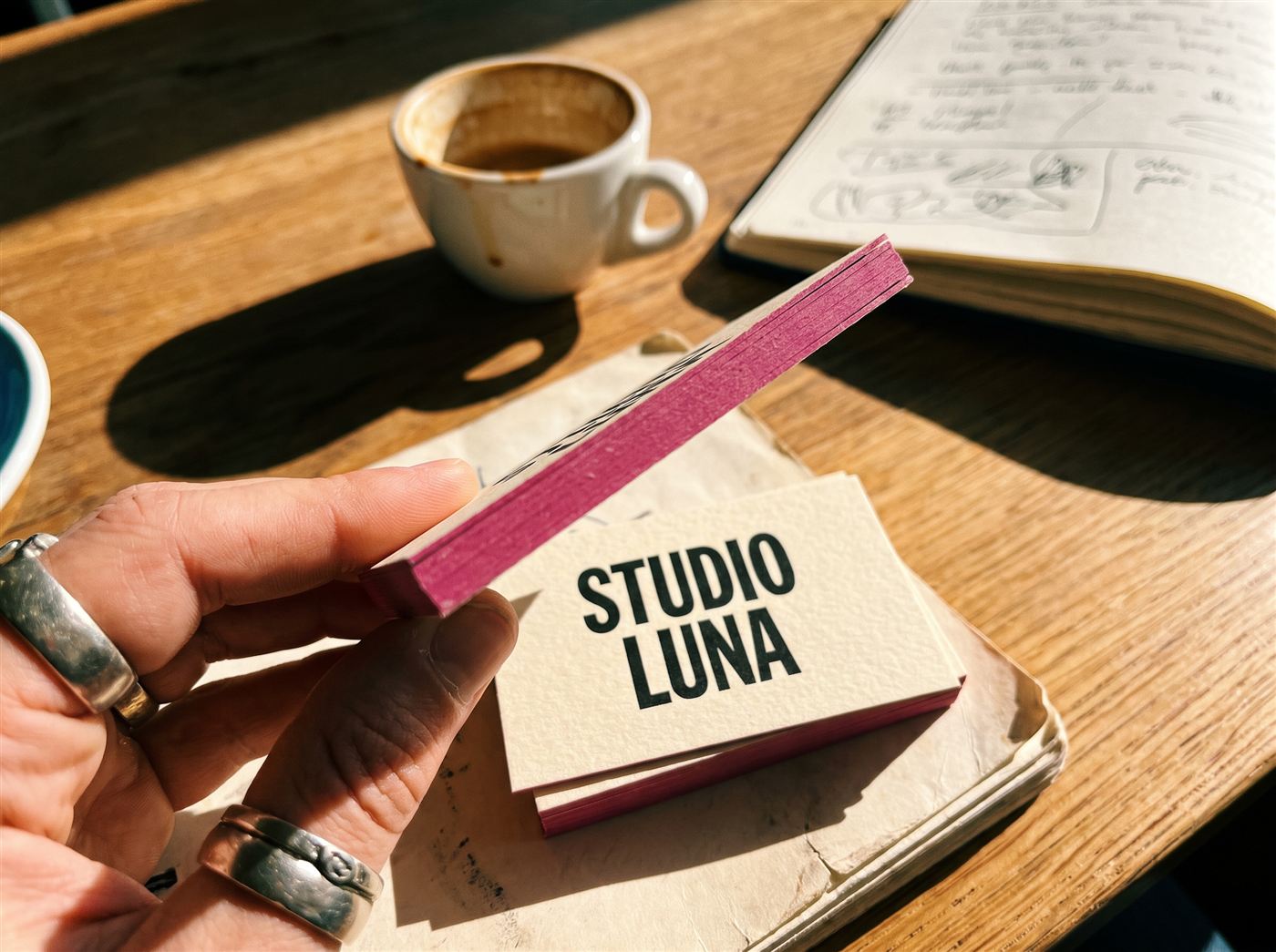 Fingers holding an ultra-thick business card edge-on showing its painted magenta edge, above a stack of cards on a café table
