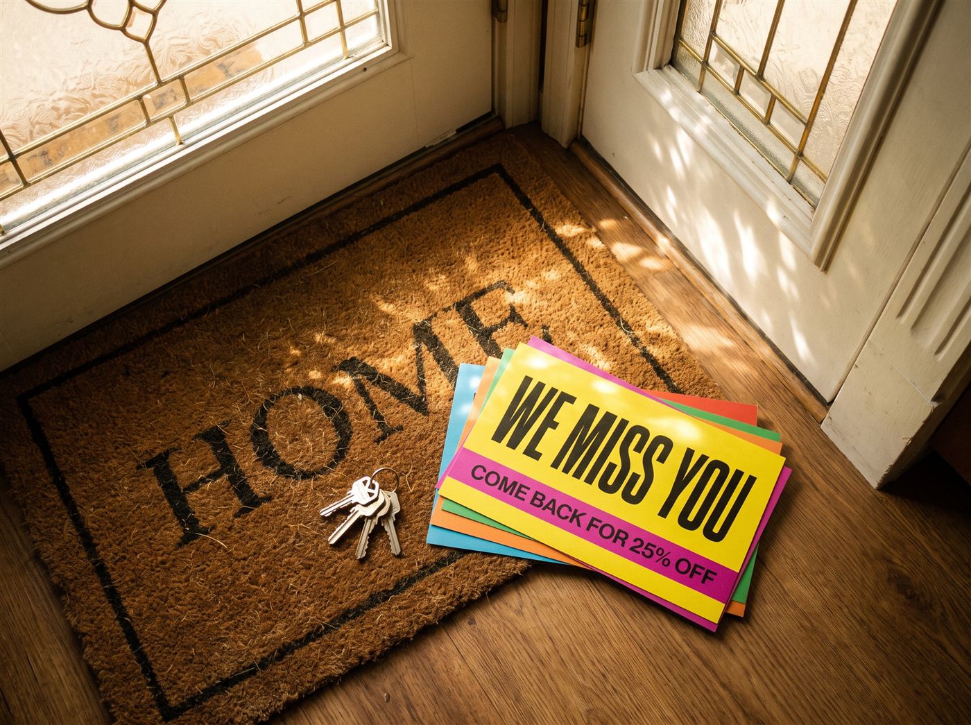 Stack of bold win-back postcards reading 'We miss you, come back for 25% off' on a doormat by a front door