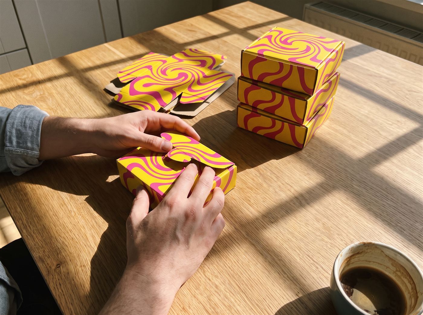 Hands folding a bright yellow product box with magenta pattern at a kitchen table next to stacked finished boxes and flat blanks